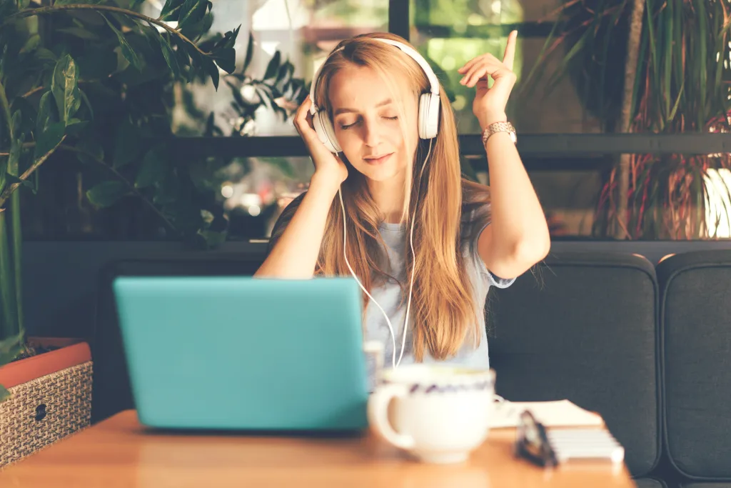 Blonde girl at a table in a cafe with headphones connected to a laptop listening to music and enjoying dancing with pleasure.